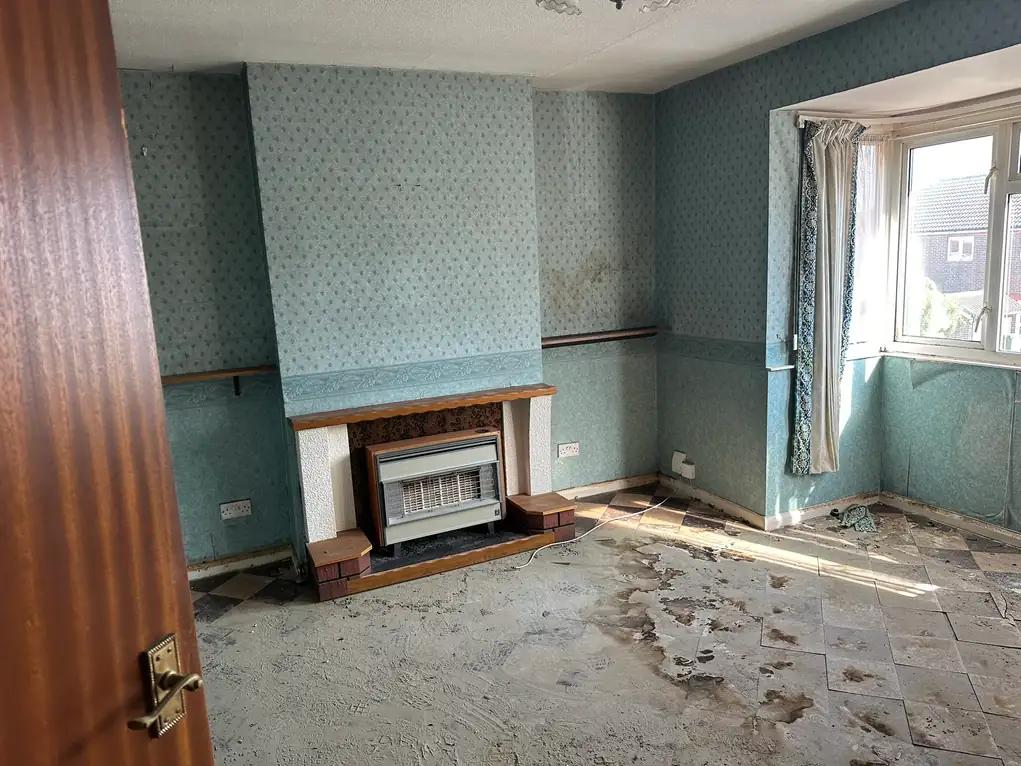 A dated living room with patterned wallpaper, dado rails and old floor tiles lifting off black adhesive, in a Sidcup house